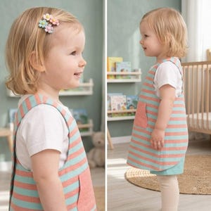 May include: A toddler wearing a white short-sleeved t-shirt under a sleeveless jumper dress featuring horizontal stripes in coral and light blue, with a solid coral patch pocket on the front and a small flower-shaped hair clip in blonde hair.