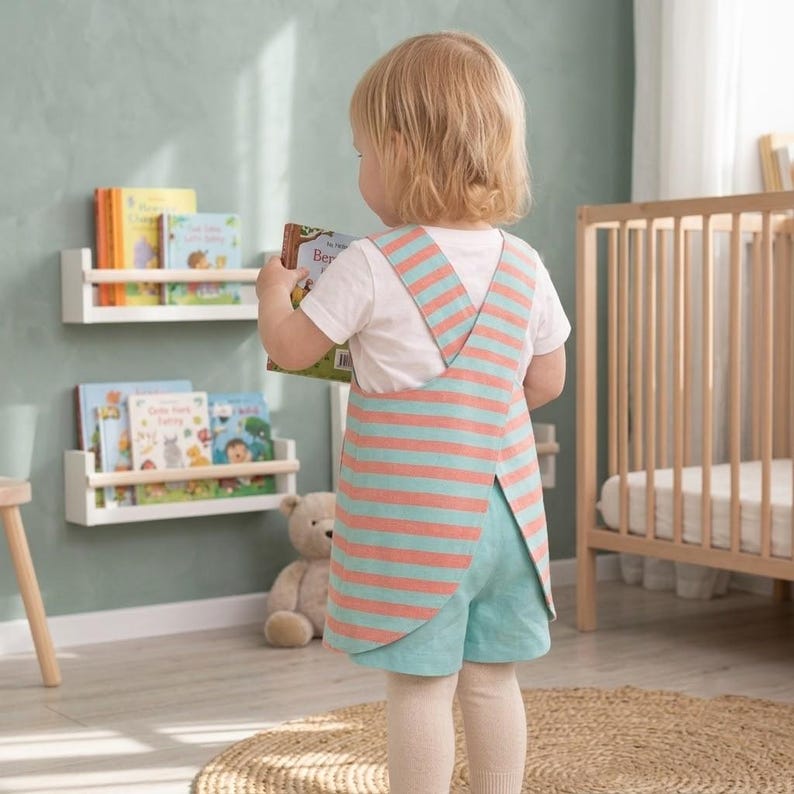 May include: A child wearing a white short-sleeved t-shirt and a sleeveless jumper with horizontal coral and light blue stripes. The jumper features a cross-back design with wide straps and a rounded hemline, paired with light blue shorts.