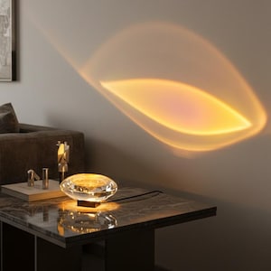 May include: A modern crystal table lamp with a unique design. The lamp projects a warm, sunset-like glow onto the wall, creating a cozy ambiance. The lamp sits on a dark marble table, with a black cord. The lamp is a decorative lighting fixture.