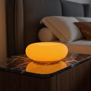 May include: A glowing, orange-yellow bedside lamp with a rounded, bowl-like shape. The lamp sits on a dark wood nightstand with a marble top. The bedroom setting includes a bed with pillows and a dark gray headboard.
