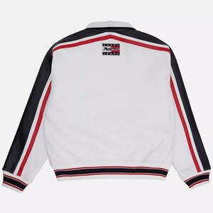 May include: White jacket with black sleeves and red stripes. The jacket has a collar and a ribbed hem with red, white, and black stripes. The back of the jacket features a small logo that says "TOMMY AVIREX JEANS".