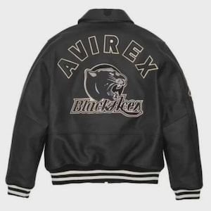 K&ouml;nnte beinhalten: Schwarze Lederjacke mit wei&szlig;-schwarz gestreiften B&uuml;ndchen und Saum. Auf der R&uuml;ckseite steht das Wort "AVIREX" &uuml;ber einer Panther-Grafik und den Worten "Black Aces". Die Jacke hat einen klassischen Kragen.