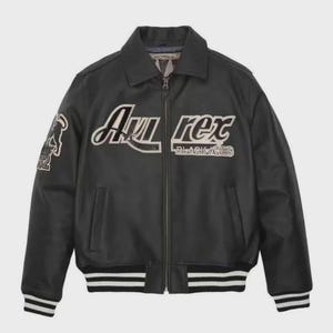K&ouml;nnte beinhalten: Schwarze Lederjacke mit Kragen, Rei&szlig;verschluss und wei&szlig; gestreiften B&uuml;ndchen und Saum. Die Jacke tr&auml;gt den Text "Avi Rex BLACKACES" in wei&szlig;er Schrift. Ein Aufn&auml;her mit einer Grafik befindet sich auf dem linken &Auml;rmel.