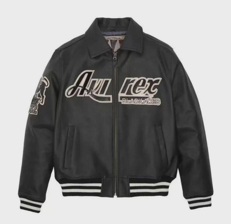 K&ouml;nnte beinhalten: Schwarze Lederjacke mit Kragen, Rei&szlig;verschluss und wei&szlig; gestreiften B&uuml;ndchen und Saum. Die Jacke tr&auml;gt den Text "Avi Rex BLACKACES" in wei&szlig;er Schrift. Ein Aufn&auml;her mit einer Grafik befindet sich auf dem linken &Auml;rmel.