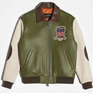 Puede incluir: Chaqueta de cuero verde oliva con mangas color crema y detalles marrones. Cuenta con un cuello marr&oacute;n, cierre de cremallera y un parche con el texto "AVIREX" y un dise&ntilde;o de bandera americana. La chaqueta tiene dobladillo y pu&ntilde;os acanalados.