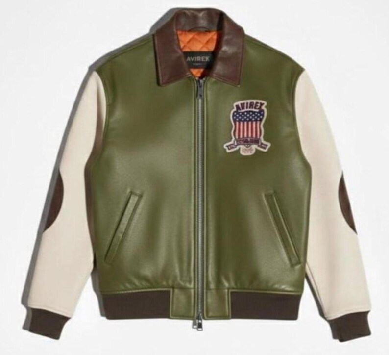 Puede incluir: Chaqueta de cuero verde oliva con mangas color crema y detalles marrones. Cuenta con un cuello marr&oacute;n, cierre de cremallera y un parche con el texto "AVIREX" y un dise&ntilde;o de bandera americana. La chaqueta tiene dobladillo y pu&ntilde;os acanalados.