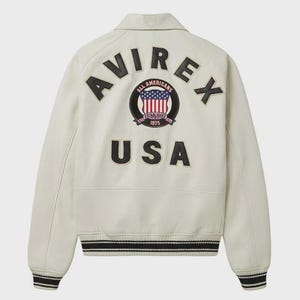 May include: Cream-colored leather jacket with black and white striped cuffs and hem. The back features the word "AVIREX" above a circular patch with the words "ALL AMERICANS ESTABLISHED 1975" and a USA flag design. Below is the word "USA".