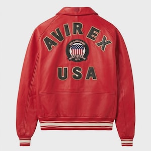 May include: A red leather jacket featuring the word "AVIREX" in black and gold lettering across the shoulders. A circular patch with "AVIREX 1975" and a USA flag design is centered below the lettering. The word "USA" is below the patch.