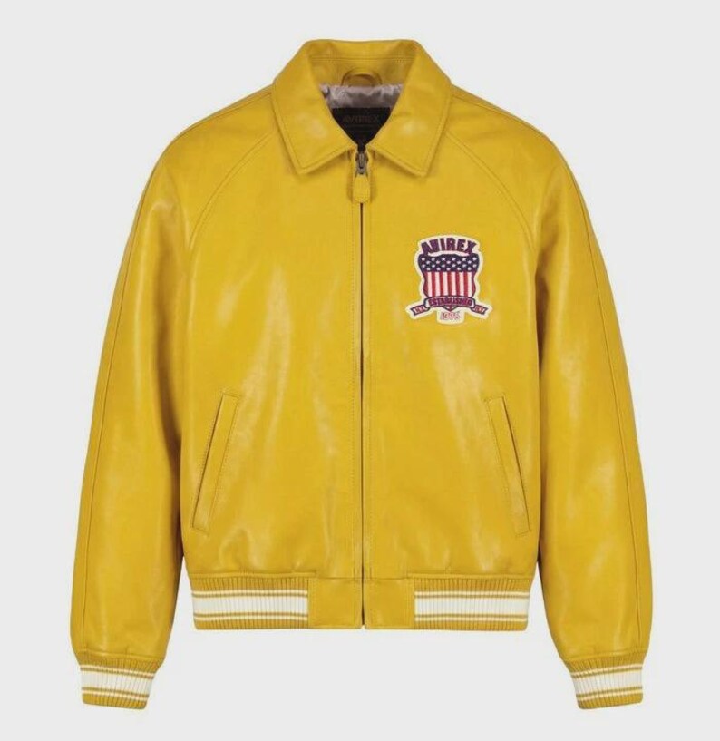 Puede incluir: Chaqueta de cuero amarillo mostaza con cierre de cremallera, cuello y pu&ntilde;os y dobladillo acanalados. Cuenta con un parche en el pecho con el texto "AVIREX" y un dise&ntilde;o con colores rojo, blanco y azul. Un estilo cl&aacute;sico de chaqueta bomber.
