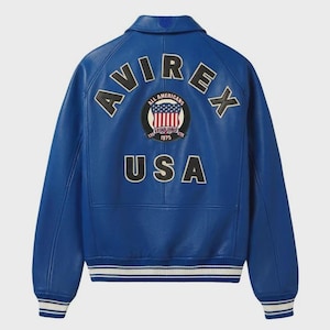 May include: A vibrant blue leather jacket with the word "AVIREX" arched across the shoulders. Below, the word "USA" is displayed. A circular patch with the words "ALL AMERICANS" and "ESTABLISHED 1975" is centered. The jacket has white and black striped cuffs.