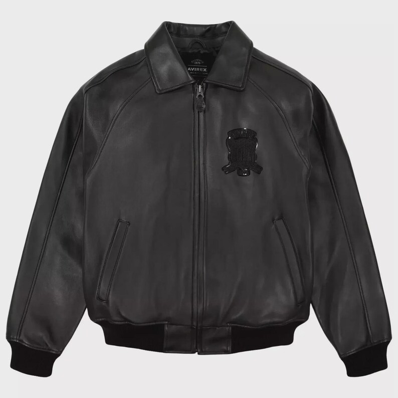 May include: A black leather jacket with a collared neckline and a zip-up front. The jacket features a black emblem on the chest and two front pockets. The cuffs and hem are ribbed. The jacket is a classic bomber style.