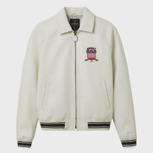 May include: Cream-colored jacket with a collared neckline and a zip-up front. Features a crest with red, white, and blue accents on the chest. The cuffs and hem have black and white striped detailing. The brand name "AVIREX" is visible.