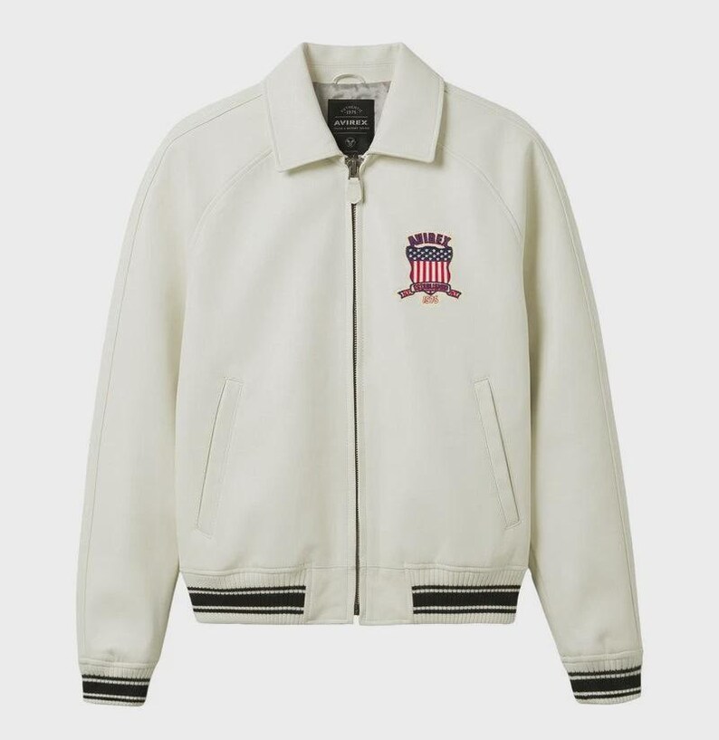 May include: Cream-colored jacket with a collared neckline and a zip-up front. Features a crest with red, white, and blue accents on the chest. The cuffs and hem have black and white striped detailing. The brand name "AVIREX" is visible.