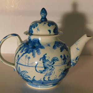 May include: A white ceramic teapot with blue hand-painted illustrations. The teapot features a round body, a curved handle, and a spout. The lid is topped with a decorative finial. The design includes a figure with a fan and landscape elements.