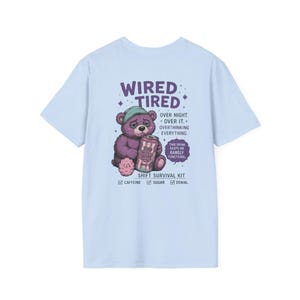 May include: Pale blue t-shirt featuring a purple bear with a green cap, holding a can of Cotton Candy soda. The shirt has the text "WIRED TIRED" and "SHIFT SURVIVAL KIT".