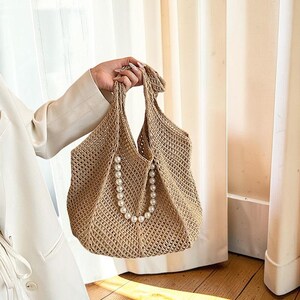 May include: A beige crocheted tote bag with a pearl necklace accent. The bag has two handles and a net-like weave. The pearl necklace hangs from the top of the bag, adding a touch of elegance. The bag is held by a person.