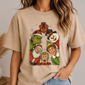 May include: A beige short-sleeve t-shirt featuring a colorful graphic print of various holiday characters including a green creature, Santa Claus, a snowman, an elf, a reindeer, and a young boy with hands on his face, surrounded by blue snowflakes.