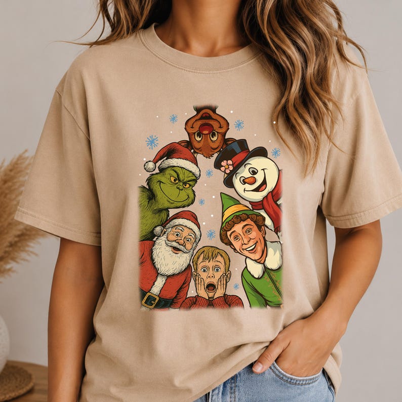 May include: A beige short-sleeve t-shirt featuring a colorful graphic print of various holiday characters including a green creature, Santa Claus, a snowman, an elf, a reindeer, and a young boy with hands on his face, surrounded by blue snowflakes.