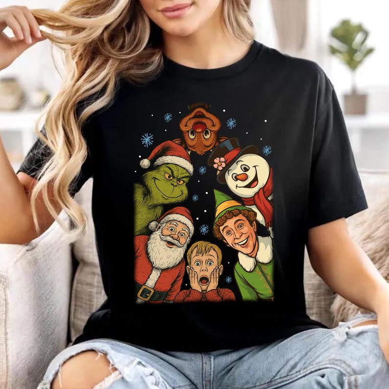 May include: A black t-shirt featuring a graphic print of various holiday characters including the Grinch, Santa Claus, a snowman, an elf, and a reindeer, surrounding a central illustration of a young boy with his hands on his face.
