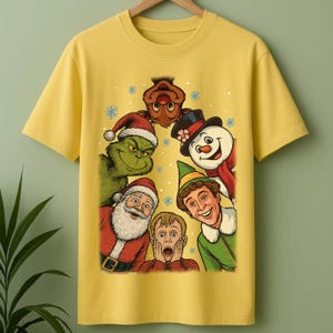 May include: A mustard yellow short-sleeve t-shirt featuring a graphic print of various holiday characters including a green creature, Santa Claus, a snowman, an elf, a reindeer, and a young boy with hands on his face, surrounded by snowflakes.