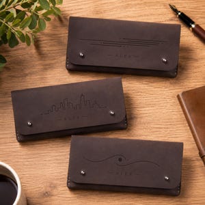 Può includere: Tre portafogli rettangolari in pelle marrone scuro con chiusure a patta fissate da due borchie in metallo color argento. Ogni portafoglio presenta un design unico inciso al laser e il nome ALEX centrato sulla patta anteriore.