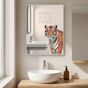 Op de afbeelding: Badkamer met een ingelijste print met een tijgerillustratie en de tekst "WASH YOUR PAWS". De print wordt weerspiegeld in een spiegel boven een witte wastafel. Een ronde witte wastafel en een houten kast staan op de voorgrond.
