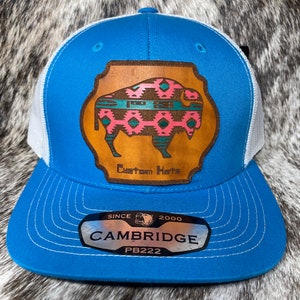 May include: A blue and white trucker hat with a brown leather patch featuring a pink and teal buffalo design. The patch says "Custom Hats". The hat also has a silver label that says "Since 2000 Cambridge PB222".