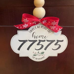 Op de afbeelding: Een kerstversiering met een rood lint met witte sneeuwvlokken. Het witte label toont de tekst "THERE'S NO PLACE LIKE home 77575 FOR THE HOLIDAYS". Opgehangen aan een natuurlijke touw en houten kralen.