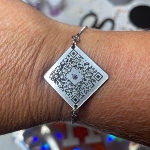 May include: A silver bracelet with a square charm featuring a black and white QR code.