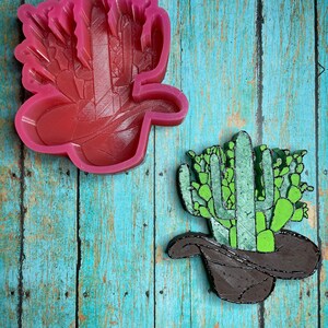 May include: A pink silicone mold of a cactus wearing a cowboy hat. The mold is next to a green and brown cactus wearing a cowboy hat.