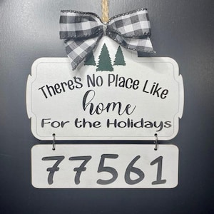 Op de afbeelding: Een wit houten bord met de tekst "There's No Place Like Home For the Holidays" en de cijfers "77561". Het bord is versierd met een zwart-wit geruite strik en groene boomafbeeldingen.