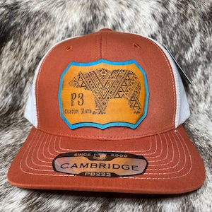 May include: A brown and white trucker hat with a leather patch featuring a bear design. The patch has a blue border and the text "P3 CUSTOM HATS". The hat has the text "CAMBRIDGE" and "PB222" on the brim.