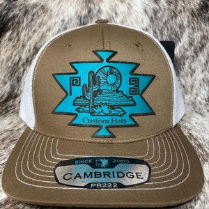May include: A brown and white trucker hat with a turquoise desert landscape design. The hat features the text "Custom Hats" and "Cambridge" with the style number PB222.