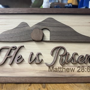 May include: Wooden sign with a light wood background and a dark wood frame. The sign features a mountain scene with an open tomb and the words "He is Risen" and "Matthew 28:6" in a decorative font.