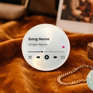 May include: A circular metallic plaque featuring a printed music player interface with the text Song Name and Singer Name, a progress bar, playback control icons, and a small pink heart icon, presented as a decorative music-themed accessory.