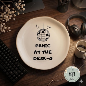 May include: A cream-colored ceramic plate with a gold rim features a black disco ball graphic and the phrase "PANIC AT THE DESK-O." The plate is on a wooden surface, with a keyboard, headphones, and a coffee cup visible.