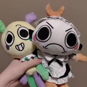 May include: Two plush dolls are held in a hand. One is yellow with green, purple, and white accents, and the other is white with black and orange accents. The dolls have large, expressive eyes and are soft toys.