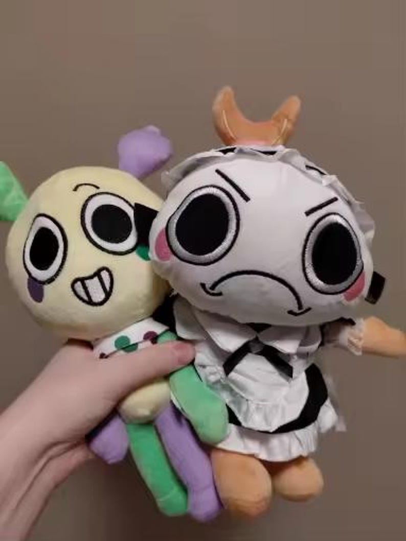 May include: Two plush dolls are held in a hand. One is yellow with green, purple, and white accents, and the other is white with black and orange accents. The dolls have large, expressive eyes and are soft toys.