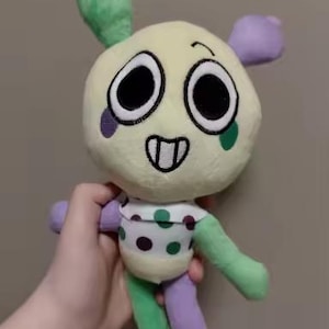 May include: A plush toy with a round, cream-colored head, large black eyes, and a smiling mouth. It has green and purple limbs, a green top, and a polka-dotted torso. The toy is held in a hand.