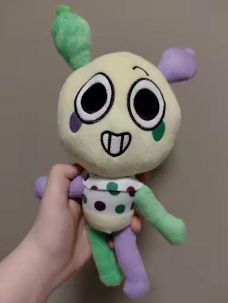 May include: A plush toy with a round, cream-colored head, large black eyes, and a smiling mouth. It has green and purple limbs, a green top, and a polka-dotted torso. The toy is held in a hand.