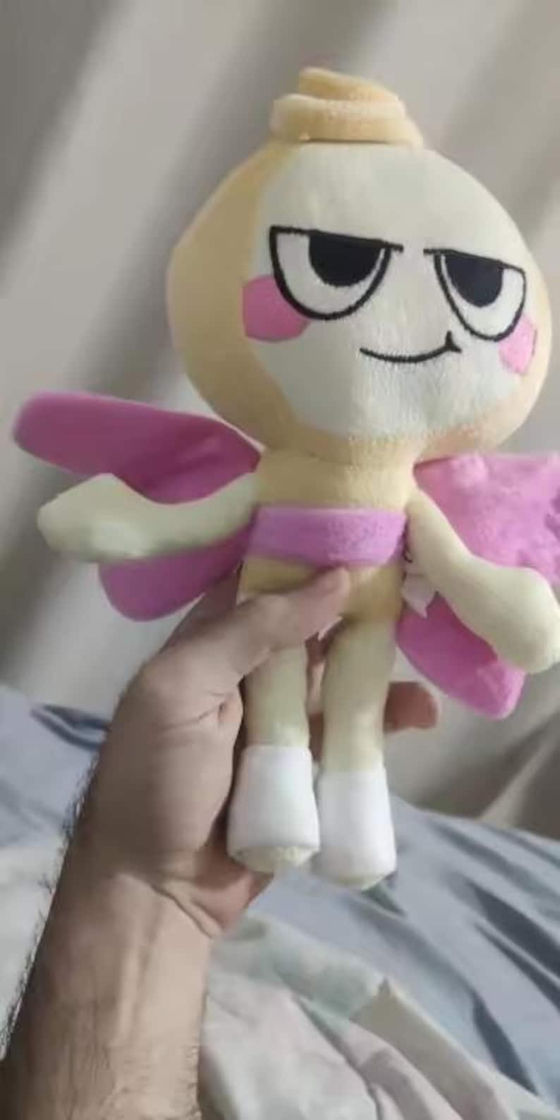 Op de afbeelding: Een knuffel van een lichtgeel personage met een rond hoofd, een kleine krul bovenop, grote zwarte ogen, een grijns en roze wangen. Het figuurtje heeft roze vleugels, een roze tailleband en witte laarsjes aan de voeten, gemaakt van zachte stof.
