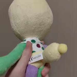 May include: A plush toy in pastel colors is held by a hand. The toy has a cream-colored head, green and purple limbs, and a polka-dot collar. A tag that says "MADE IN CHINA" is visible.