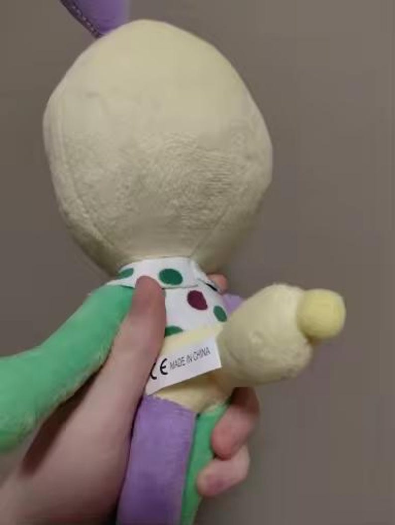 May include: A plush toy in pastel colors is held by a hand. The toy has a cream-colored head, green and purple limbs, and a polka-dot collar. A tag that says "MADE IN CHINA" is visible.