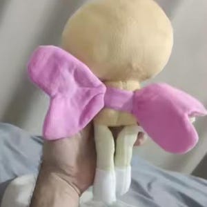 Op de afbeelding: Een knuffel met een rond, lichtgeel hoofd met een klein plukje bovenop, een paar zachte roze vleugels aan de achterkant en twee lichtgele benen die eindigen in witte voeten. Het speelgoed is gemaakt van een zacht, pluizig materiaal.