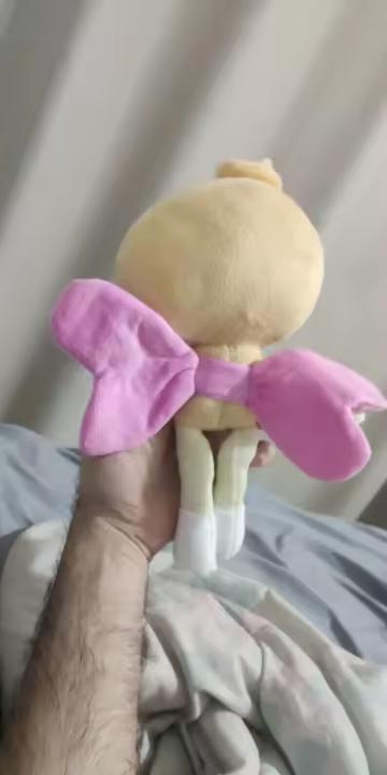 Op de afbeelding: Een knuffel met een rond, lichtgeel hoofd met een klein plukje bovenop, een paar zachte roze vleugels aan de achterkant en twee lichtgele benen die eindigen in witte voeten. Het speelgoed is gemaakt van een zacht, pluizig materiaal.