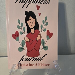 May include: A pink journal titled "Happiness Journal" with a graphic of a person hugging themself. The cover also features red hearts, green leaves, and the author's name, Christine A Fisher, in black script.