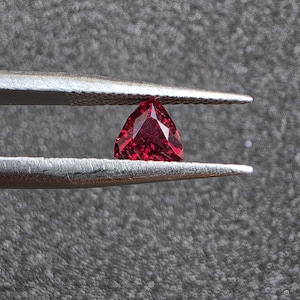 May include: A vibrant, deep red gemstone cut into a triangular shape with multiple facets. The gemstone is held securely between the two metal tips of a pair of silver-toned tweezers, showcasing its brilliant clarity and rich crimson color.