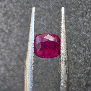 Può includere: Una pietra preziosa di rubino taglio cuscino, di colore rosso intenso e superficie sfaccettata, tenuta tra le due punte metalliche di una pinzetta color argento. La gemma mostra chiarezza interna e riflessi di luce sulle faccette.
