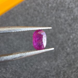Può includere: Una gemma color magenta dal taglio ovale tenuta da una pinzetta in metallo color argento. La pietra presenta una superficie sfaccettata che riflette la luce, mostrando una tonalità rosa-viola intenso con inclusioni interne visibili.