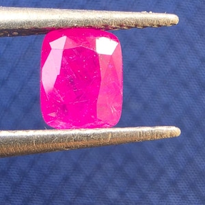 May include: A vibrant, cushion-cut hot pink gemstone held between the metal tips of a pair of tweezers. The stone features multiple facets that reflect light, showcasing a deep, saturated magenta hue with visible internal inclusions and clarity.